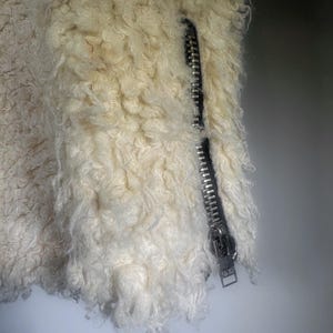 Puede incluir: Primer plano de una chaqueta color crema con cremallera plateada. La chaqueta tiene una textura rizada y esponjosa. La cremallera est&aacute; cerrada, lo que sugiere un material c&aacute;lido, posiblemente de borrego.