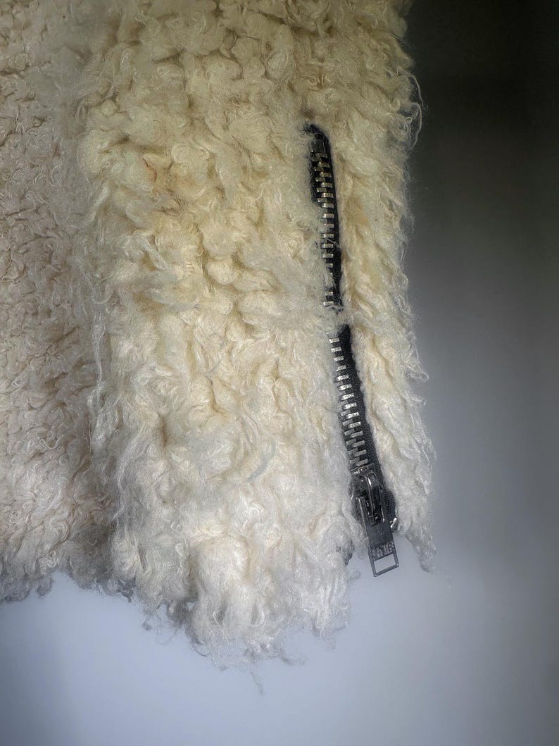 Puede incluir: Primer plano de una chaqueta color crema con cremallera plateada. La chaqueta tiene una textura rizada y esponjosa. La cremallera est&aacute; cerrada, lo que sugiere un material c&aacute;lido, posiblemente de borrego.