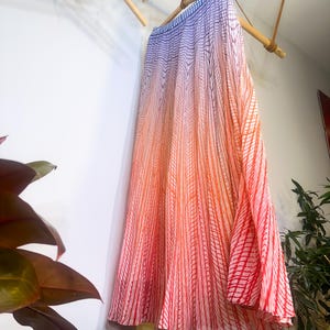 May include: A long, flowing skirt with a gradient design, transitioning from blue at the top to orange and red at the bottom. The skirt features vertical striped patterns and is hanging from a bamboo rod.