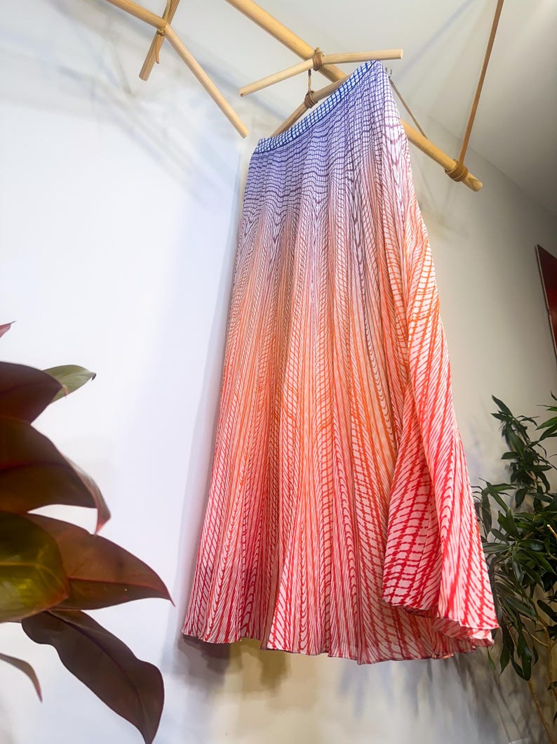 May include: A long, flowing skirt with a gradient design, transitioning from blue at the top to orange and red at the bottom. The skirt features vertical striped patterns and is hanging from a bamboo rod.