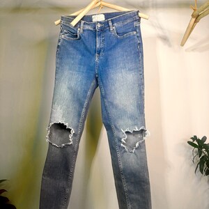Op de afbeelding: Blauwe denim jeans met distressed knie-uitsnijdingen die aan een houten hanger hangen. De jeans hebben een vervaagde wassing en een slanke pasvorm. De denim is van gemiddeld gewicht, geschikt voor casual wear.