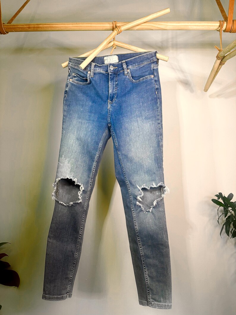 Op de afbeelding: Blauwe denim jeans met distressed knie-uitsnijdingen die aan een houten hanger hangen. De jeans hebben een vervaagde wassing en een slanke pasvorm. De denim is van gemiddeld gewicht, geschikt voor casual wear.