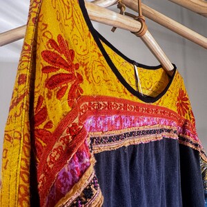 Puede incluir: Una blusa negra y azul bordada con ribetes amarillos, rojos y rosas. La blusa tiene un escote redondo y est&aacute; colgada de una percha de madera.