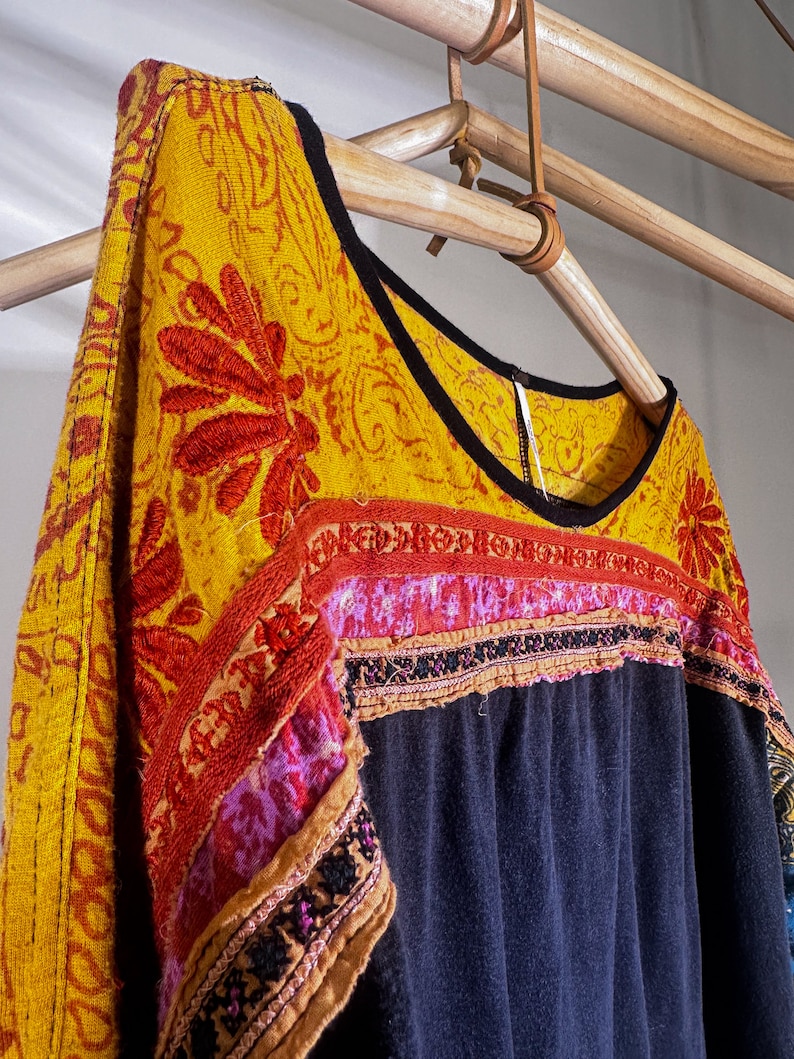 Puede incluir: Una blusa negra y azul bordada con ribetes amarillos, rojos y rosas. La blusa tiene un escote redondo y est&aacute; colgada de una percha de madera.