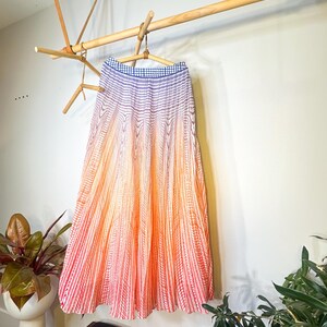 May include: A long, pleated skirt with a gradient design, transitioning from blue at the waistband to orange and red at the hem. The skirt is hanging on a wooden hanger, suspended from a wooden rack. The skirt's fabric has vertical striped patterns.