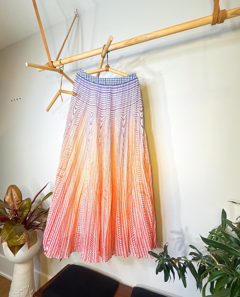 May include: A long, pleated skirt with a gradient design, transitioning from blue at the waistband to orange and red at the hem. The skirt is hanging on a wooden hanger, suspended from a wooden rack. The skirt's fabric has vertical striped patterns.