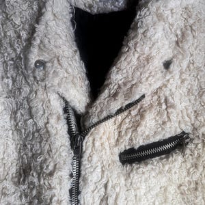 Puede incluir: Chaqueta texturizada color crema con cremallera negra y un peque&ntilde;o bolsillo negro con cremallera. La chaqueta tiene una textura rizada y difusa y una peque&ntilde;a etiqueta que dice "UNIF x Urban Outfitters".
