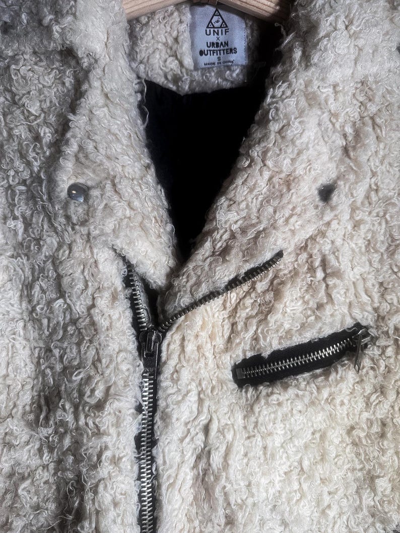 Puede incluir: Chaqueta texturizada color crema con cremallera negra y un peque&ntilde;o bolsillo negro con cremallera. La chaqueta tiene una textura rizada y difusa y una peque&ntilde;a etiqueta que dice "UNIF x Urban Outfitters".