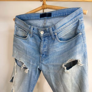 May include: Pale blue denim jeans with distressed detailing, featuring large rips on the legs. The jeans are displayed on a wooden hanger, highlighting the button-fly closure and classic five-pocket design. The denim has a faded wash.
