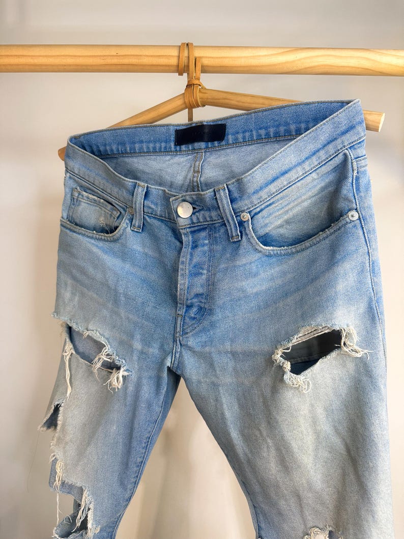May include: Pale blue denim jeans with distressed detailing, featuring large rips on the legs. The jeans are displayed on a wooden hanger, highlighting the button-fly closure and classic five-pocket design. The denim has a faded wash.