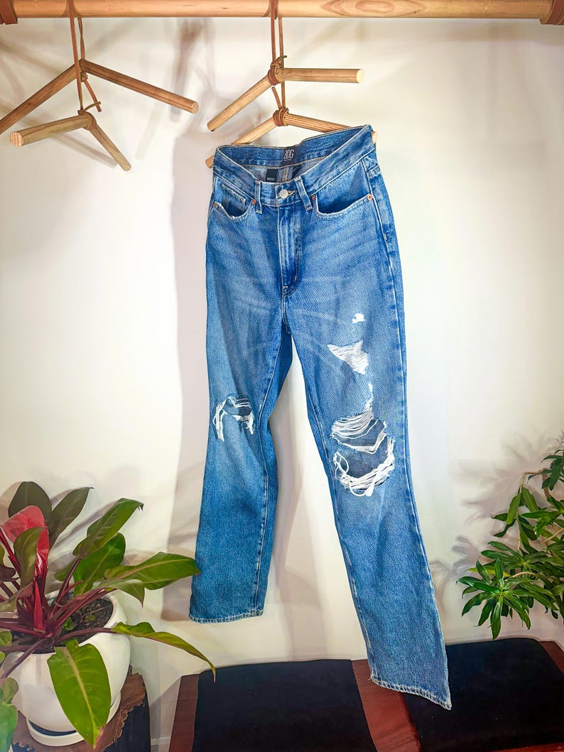 Op de afbeelding: Een lichtblauwe spijkerbroek met distressed details, hangend aan een houten hanger.