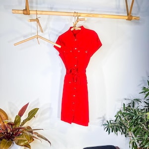 May include: A bright red dress with short sleeves and a button-down front hangs on a wooden hanger. The dress features a matching belt and small pockets. The dress is displayed against a white wall with plants in the foreground.