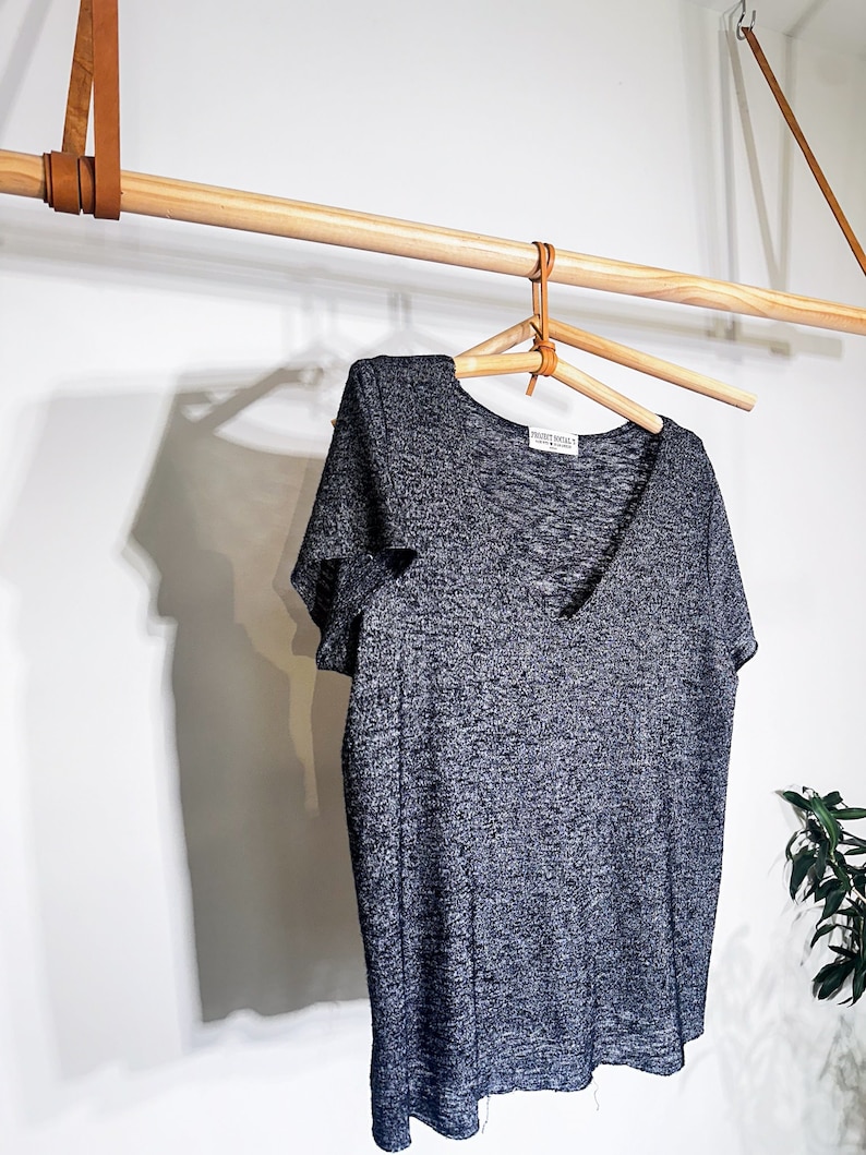May include: A gray, short-sleeved, V-neck t-shirt hanging on a wooden rack with a leather strap. The shirt has a textured, slightly speckled fabric. The shirt has a small white tag with the text "FRONT ROW" on it.