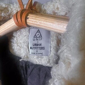 Puede incluir: Primer plano de una chaqueta crema texturizada con forro negro. La chaqueta est&aacute; colgada de una percha de madera con lazos de cuero marr&oacute;n. Una etiqueta dice "UNIF X Urban Outfitters" y "Made in China."