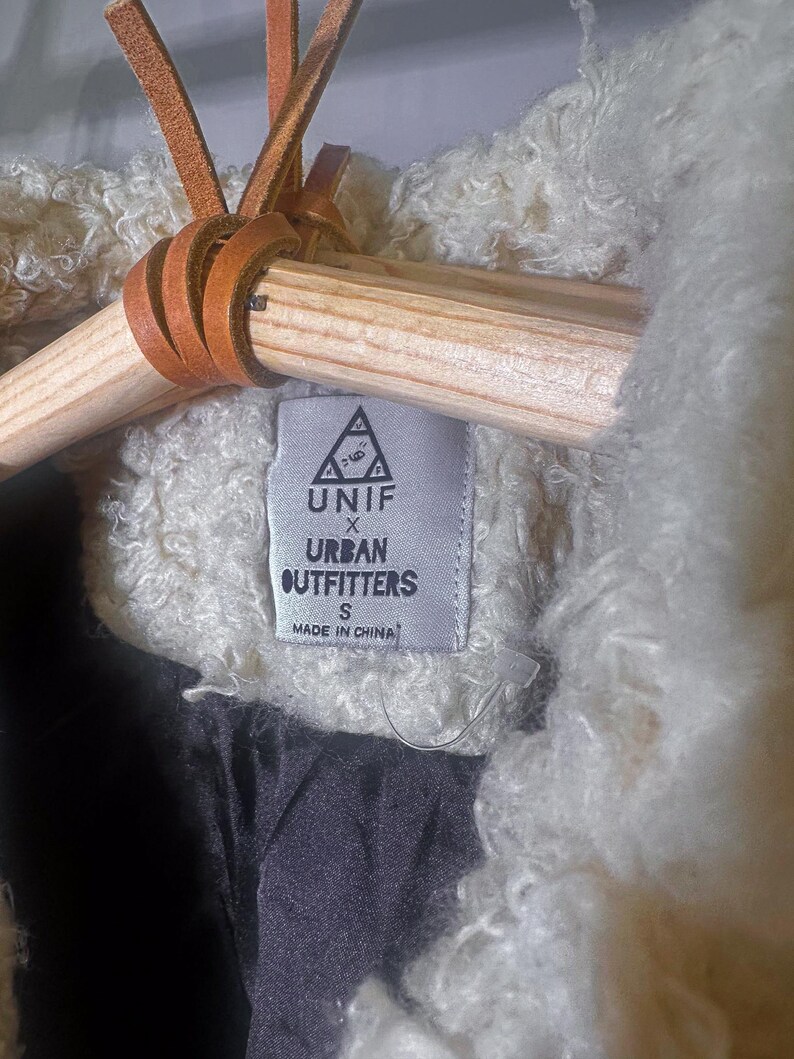 Puede incluir: Primer plano de una chaqueta crema texturizada con forro negro. La chaqueta est&aacute; colgada de una percha de madera con lazos de cuero marr&oacute;n. Una etiqueta dice "UNIF X Urban Outfitters" y "Made in China."