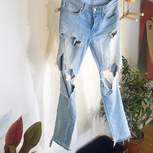 May include: Pale blue denim jeans with distressed detailing, featuring large rips on the legs. The jeans are displayed on a wooden rack, highlighting their design. They have a button fly and classic five-pocket styling.