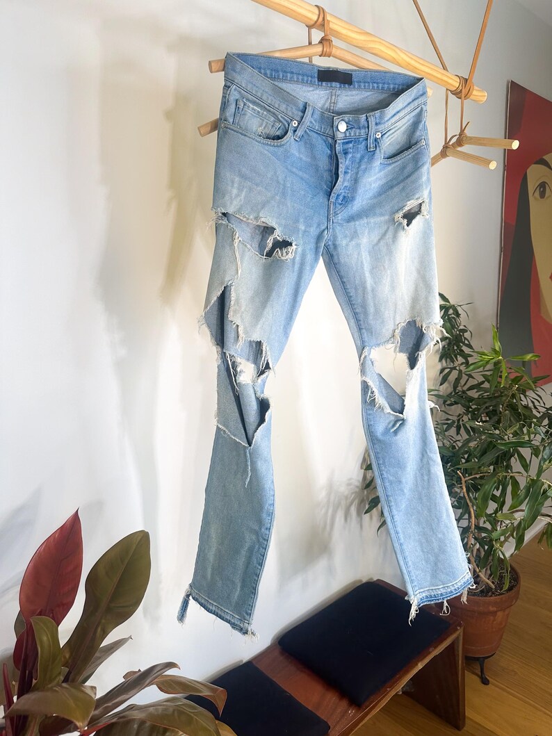May include: Pale blue denim jeans with distressed detailing, featuring large rips on the legs. The jeans are displayed on a wooden rack, highlighting their design. They have a button fly and classic five-pocket styling.