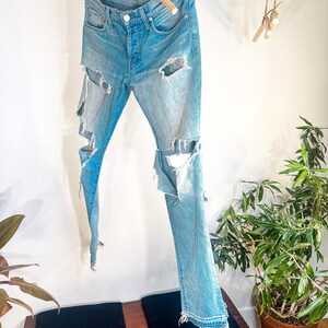 May include: Light blue denim jeans with distressed detailing, featuring large rips on the legs. The jeans are suspended on a wooden hanger. The jeans have a button fly and classic five-pocket design. The jeans are hanging against a white wall.