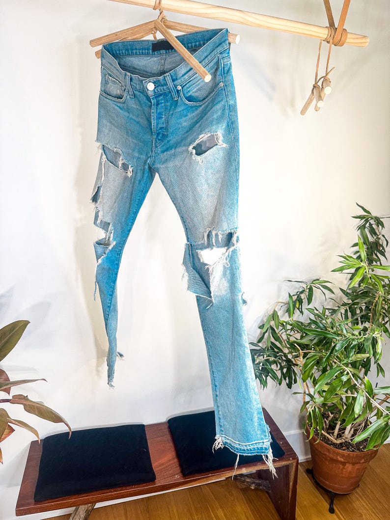 May include: Light blue denim jeans with distressed detailing, featuring large rips on the legs. The jeans are suspended on a wooden hanger. The jeans have a button fly and classic five-pocket design. The jeans are hanging against a white wall.
