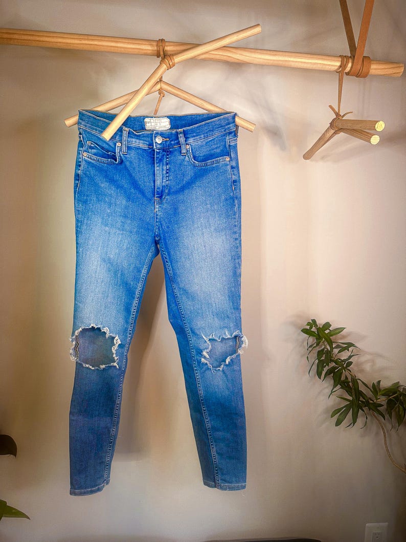 May include: Light wash blue denim jeans with distressed knees hanging on a wooden clothes hanger. The jeans are a skinny fit and have a mid-rise waist.  The brand label is visible on the back waistband.