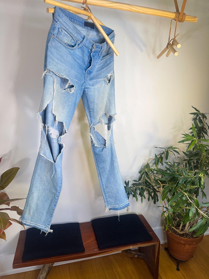 May include: Light blue denim jeans with distressed details, hanging from a wooden rod. The jeans have large rips on the front of the legs. The jeans are hanging above a wooden bench with two black cushions.