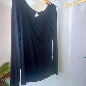 Puede incluir: Una camisa negra de manga larga con cuello en V y detalle de envoltura. La camisa est&aacute; colgada en una percha de madera.