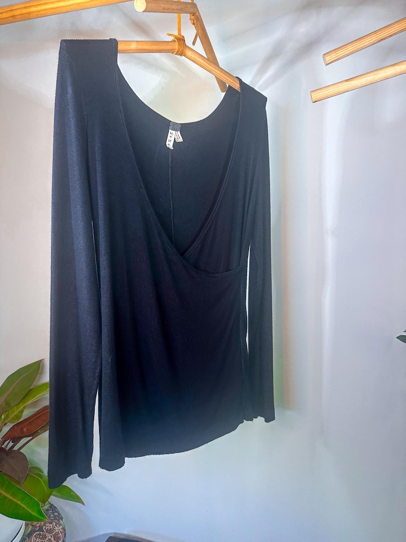 Puede incluir: Una camisa negra de manga larga con cuello en V y detalle de envoltura. La camisa est&aacute; colgada en una percha de madera.