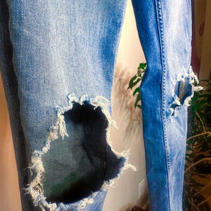Op de afbeelding: Lichtblauwe denim jeans met distressed details, met grote, gerafelde gaten bij de knie&euml;n. De jeans hebben een klassiek ontwerp met vijf zakken en een vervaagde wassing, waardoor een versleten vintage esthetiek ontstaat. De stof vertoont tekenen van slijtage.