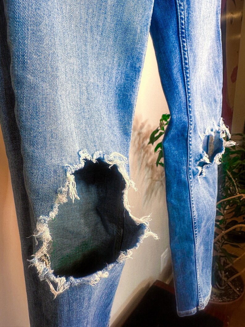 Op de afbeelding: Lichtblauwe denim jeans met distressed details, met grote, gerafelde gaten bij de knie&euml;n. De jeans hebben een klassiek ontwerp met vijf zakken en een vervaagde wassing, waardoor een versleten vintage esthetiek ontstaat. De stof vertoont tekenen van slijtage.