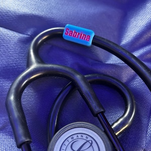 Könnte beinhalten: Ein schwarzes Stethoskop mit einem blauen Namensschild, auf dem "Sabrina" steht, und einem silbernen Littmann-Logo.