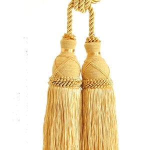 Op de afbeelding: Goudkleurige gordijn tiebacks met een decoratief kwastontwerp. De tiebacks hebben een gedraaide touwtop, een cilindrisch lichaam omwikkeld met gouden draad en een kralenrand. De kwasten zijn gemaakt van lange, vloeiende strengen.