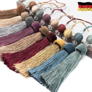 Op de afbeelding: Assortiment van kleurrijke kwastversiering met decoratieve kralen. De kwasten zijn gemaakt van zachte, zijdeachtige stof en zijn verkrijgbaar in verschillende kleuren, waaronder grijs, bordeauxrood, beige, rood en blauw.