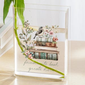 May include: Clear rectangular vase with a floral and bird design. The vase features a stack of books, a bird, and the text "favorite book quote here." Green bamboo and leaves are inside the vase, which is filled with water.