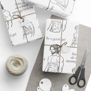 May include: White gift wrap featuring a black line drawing of a muscular man. The paper is tied with twine and a star-shaped tag. Text on the wrap includes "Be a good girl" and "Have an absolutely amazing birthday."
