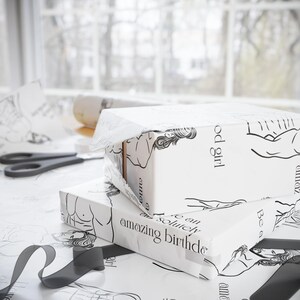 May include: Two gift boxes wrapped in white paper with black line art illustrations of figures and text. The wrapping paper has the text "amazing birthday" and "good girl". A pair of scissors and a roll of paper are also visible.