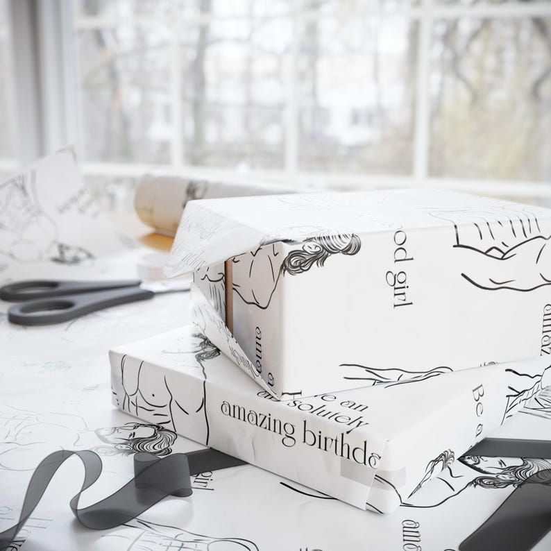 May include: Two gift boxes wrapped in white paper with black line art illustrations of figures and text. The wrapping paper has the text "amazing birthday" and "good girl". A pair of scissors and a roll of paper are also visible.
