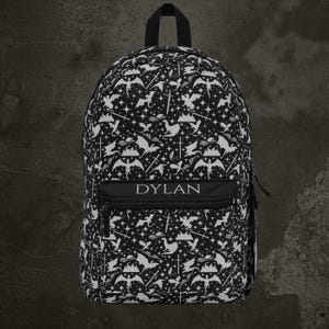 May include: Black and white backpack with a repeating pattern of bats, stars, and swords. The backpack has a black zipper pocket with the name "DYLAN" in white letters. It has a black handle and black zippers.