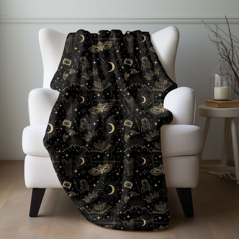 Fantasy inspired blanket with a celestial print in tan on a black blanket, the pattern includes images of moons, masks, bat wings, books, harp, fae and stars for fans of acotar and sarah j maas