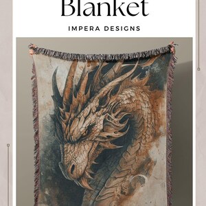 May include: A woven throw blanket featuring a detailed illustration of a dragon's head. The blanket is brown, beige, and gray with a textured woven design. The text "Dragon Woven Blanket" and "Impera Designs" is printed on the white background above the blanket. The text "For Fans of Fantasy and Castlecore" is printed on the white background below the blanket.