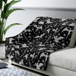 May include: A black and grey throw blanket featuring a repeating pattern of dragons, stars, and swords. The blanket is soft and cosy, ideal for snuggling up on the sofa.