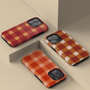 Puede incluir: Tres fundas para teléfono con un patrón de cuadros en tonos rojos, naranjas y marrones. Las fundas tienen un acabado brillante y un borde negro. Se presentan sobre una superficie de color neutro.