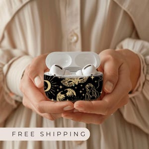 May include: A black and gold Airpods case with a celestial design featuring stars, moons, a mask, and a woman's face.
