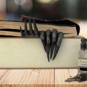 Könnte beinhalten: Ein schwarzes Buch mit einem Paar schwarzer, krallenbewehrter Hände, die es offen halten. Eine kleine Flasche mit einem Etikett, auf dem "Inchiostro appena" steht, befindet sich auf einer Holzoberfläche neben einer schwarzen und silbernen Feder.