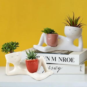 May include: Two white ceramic planters shaped like reclining figures, each holding a small potted succulent. One planter has a red pot, the other a pink pot. The planters are on a white surface with a yellow background. The planters have closed eyes and smiling faces.