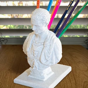 May include: A white bust pencil holder featuring a classical figure with colorful pencils. The bust is on a rectangular base. The pencils are various colors, including blue, pink, and green.