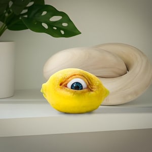 Può includere: Un limone giallo con un occhio blu dipinto su di esso, seduto su uno scaffale bianco accanto a una scultura in legno marrone chiaro.