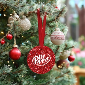 May include: A red glittery Dr. Pepper Christmas ornament with the brand's logo in white, hanging from a red ribbon. The ornament is round and surrounded by other Christmas ornaments on a decorated tree.