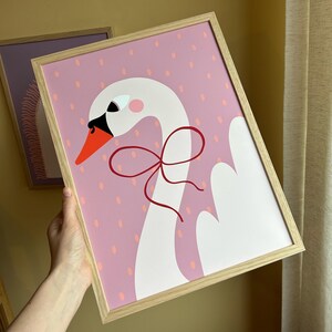 May include: A white swan with a red bow on its neck, against a pink background with white polka dots.
