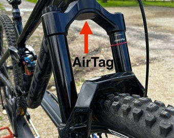 Può includere: Una mountain bike nera con una forcella nera e un manubrio nero. La forcella ha un tappo nero in cima con una freccia che punta verso di esso e il testo "AirTag" sotto la freccia. La bicicletta è su un portabiciclette.