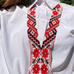 May include: A beaded necklace with a V-shaped design. The necklace features a pattern of red flowers, black and white geometric shapes, and red fringe. The necklace is worn over a white collared shirt.